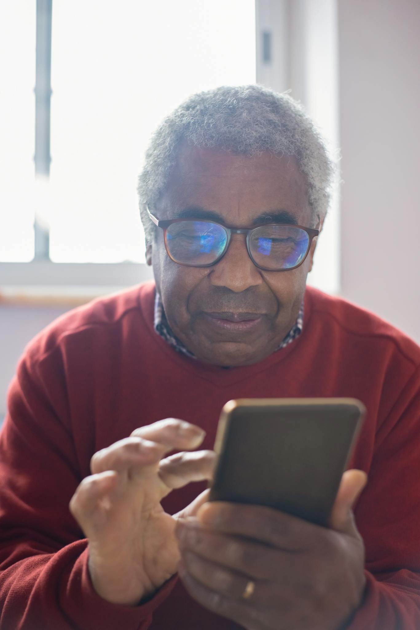 Elderly man with gray hair using a smartphone, sitting indoors with eyeglasses.